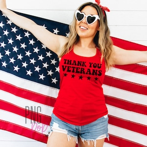 May include: A woman wearing a red tank top with the text "Thank You Veterans" and five stars printed on it. She is holding an American flag in front of a white wood background.