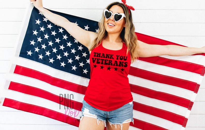 May include: A woman wearing a red tank top with the text "Thank You Veterans" and five stars printed on it. She is holding an American flag in front of a white wood background.