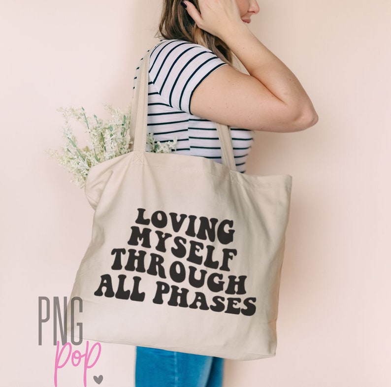 May include: A beige canvas tote bag with black text that reads "Loving Myself Through All Phases".
