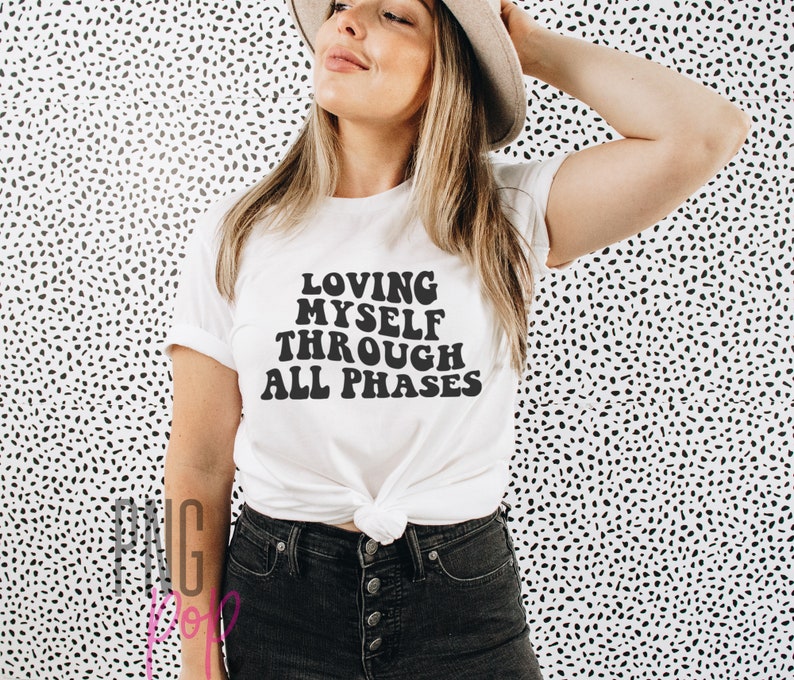 May include: White t-shirt with black text that reads "Loving Myself Through All Phases" in a retro font.