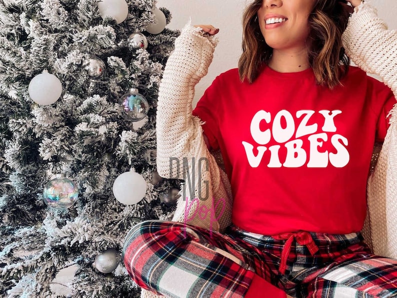 Puede incluir: Una mujer lleva una camiseta roja con el texto "Cozy Vibes" en blanco con fuente retro. Lleva pantalones de pijama a cuadros y una chaqueta blanca. Est&aacute; de pie junto a un &aacute;rbol de Navidad.