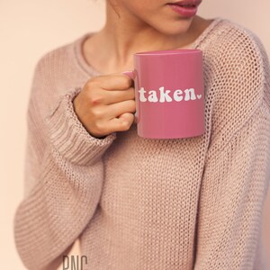 May include: A pink ceramic mug with a white heart and the word "taken" in white letters. The mug is being held by a person wearing a pink sweater.