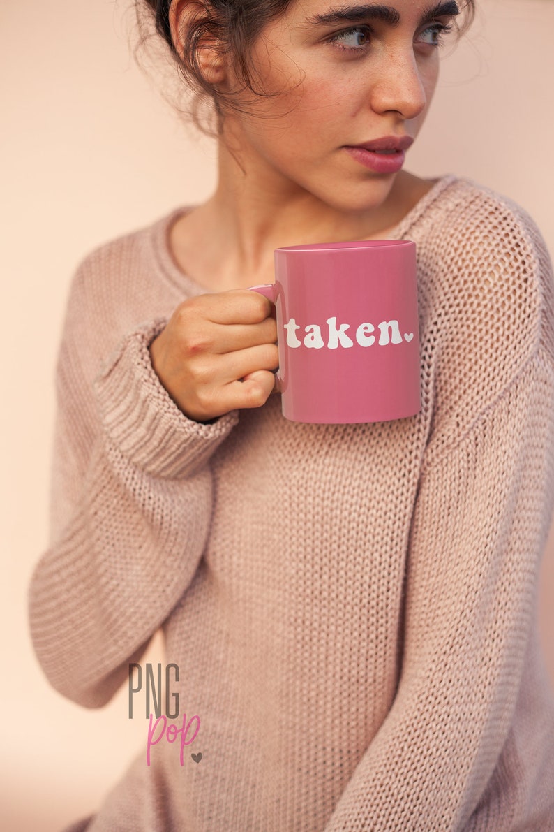 May include: A pink ceramic mug with a white heart and the word "taken" in white letters. The mug is being held by a person wearing a pink sweater.