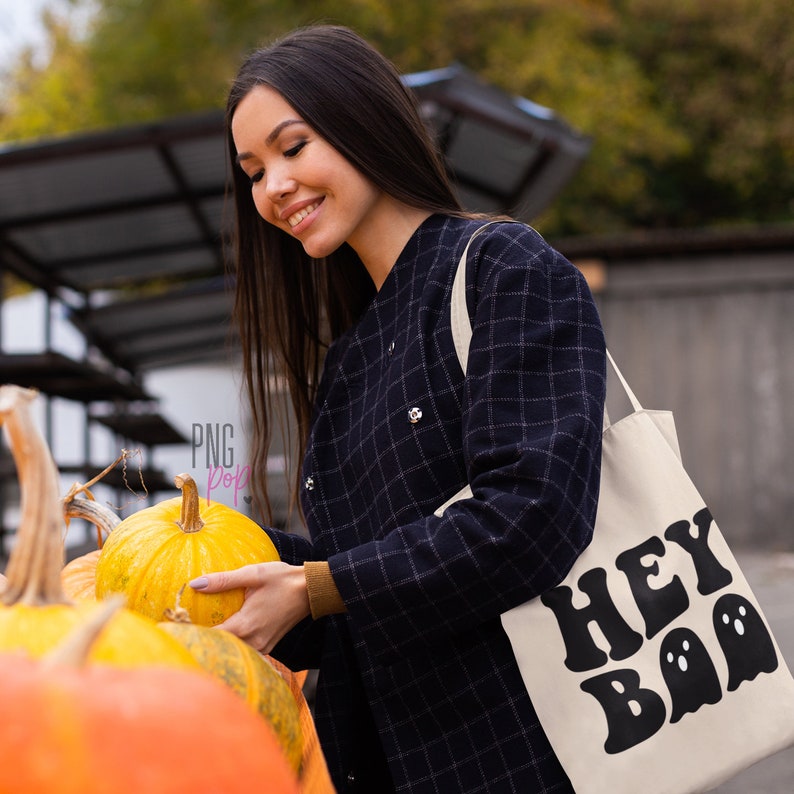 May include: A woman in a black and white checkered jacket holds a white tote bag with the text "HEY BOO" in black with ghost silhouettes. She is holding a yellow pumpkin.
