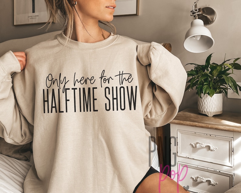 K&ouml;nnte beinhalten: Ein beiger Pullover mit dem Aufdruck "Only here for the HALFTIME SHOW" in Schwarz.