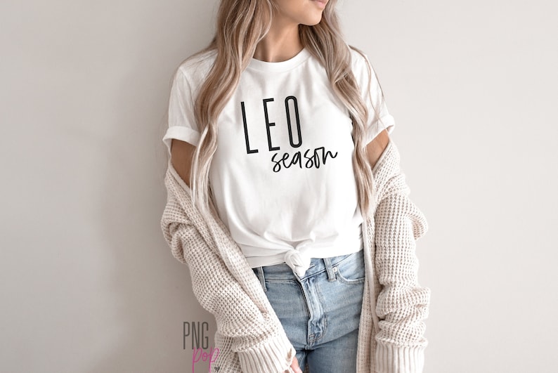 May include: White t-shirt with black text that reads "LEO season". The person wearing the shirt is also wearing a light brown cardigan and blue jeans.