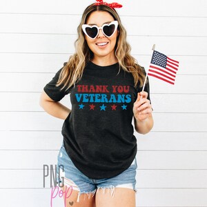 May include: A woman wearing a black t-shirt with the text "Thank You Veterans" in red and blue lettering with five white stars underneath. She is holding a small American flag in her right hand.