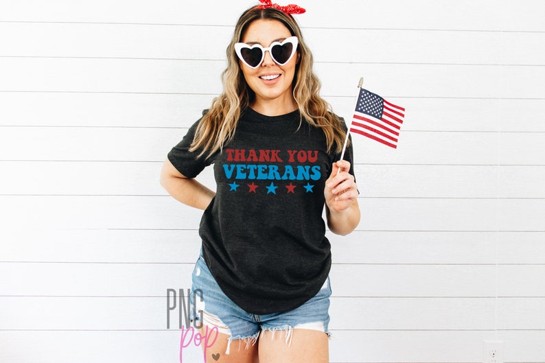 May include: A woman wearing a black t-shirt with the text "Thank You Veterans" in red and blue lettering with five white stars underneath. She is holding a small American flag in her right hand.