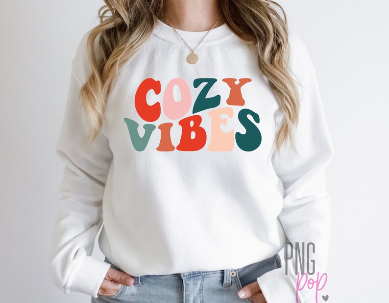 Puede incluir: Sudadera blanca con el texto "Cozy Vibes" en letras retro de colores.
