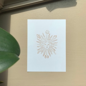 K&ouml;nnte beinhalten: Eine Linienzeichnung einer Sonne mit einem Gesicht, umgeben von stilisierten Blumen und Bl&auml;ttern. Die Zeichnung ist in Gold auf wei&szlig;em Hintergrund.