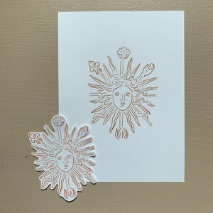 K&ouml;nnte beinhalten: Ein Aufkleber und ein Druck einer Sonne mit einem Gesicht, umgeben von Strahlen und stilisierten Blumen. Das Design ist in brauner Farbe auf wei&szlig;em Hintergrund.