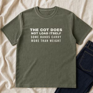 May include: An olive green short-sleeved t-shirt featuring white text printed on the chest that reads: THE COT DOES NOT LOAD ITSELF SOME HANDS CARRY MORE THAN WEIGHT. The shirt has a crew neckline and a standard fit, displayed flat on a fabric surface.