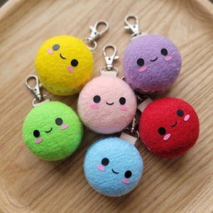May include: A collection of six round, felted macaron-shaped keychains in yellow, purple, green, pink, red, and blue. Each keychain features a smiling face with black circular eyes and small pink cheeks, attached to a silver metal lobster clasp.