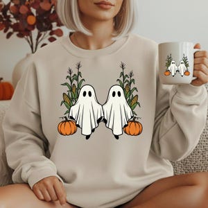 Peut inclure: Un sweat-shirt à col rond beige orné d'un motif représentant deux fantômes blancs entourés de pieds de maïs et de citrouilles orange. Une tasse en céramique blanche assortie est tenue sur le côté avec la même illustration de fantômes.