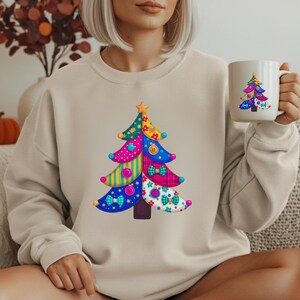Peut inclure: Un sweat-shirt beige à col rond orné d'un motif de sapin de Noël coloré façon patchwork, agrémenté de boutons, de nœuds et de motifs variés. Une tasse en céramique blanche assortie présente le même sapin de Noël multicolore.