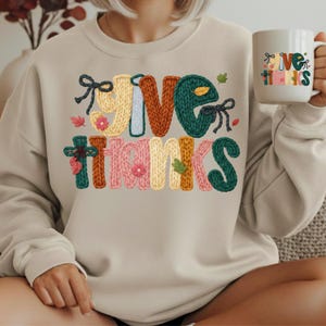 Peut inclure: Un sweat-shirt beige à col rond arborant un motif avec le texte "GIVE THANKS" dans une police texturée imitant le tricot. Les lettres sont colorées en jaune, orange, vert et rose, agrémentées de petits nœuds noirs, de fleurs et de feuilles.