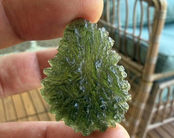 May include: A translucent, olive-green moldavite specimen with a highly textured, irregular surface featuring deep ridges and sharp, jagged edges. The stone displays a vibrant, forest-green hue with varying levels of light transmission throughout.
