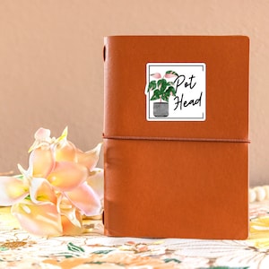 May include: A brown leather journal with a sticker on the front. The sticker features a watercolor illustration of a potted plant with the text "Pot Head".