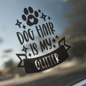May include: Black vinyl car decal with the text "DOG HAIR IS MY GLITTER" in a playful font, featuring a paw print and star accents. The decal is applied to a car window, showcasing a fun, pet-themed accessory.
