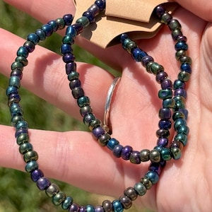 May include: Two iridescent beaded necklaces in shades of blue, green, and purple. The beads are small and round, strung together to form a delicate necklace. The necklaces are displayed on a brown tag.