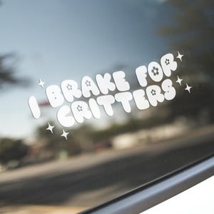 May include: White vinyl car decal with the text "I brake for critters" in a playful, rounded font. The decal is adorned with star accents, and is affixed to a car window, with a blurred background.