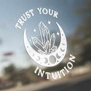 May include: A white vinyl decal applied to a car window featuring a crescent moon, a cluster of crystals, and the text "TRUST YOUR INTUITION" arranged in a circular pattern around the central graphic elements.