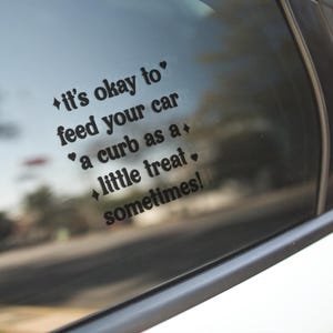 May include: A black vinyl decal applied to a car window featuring the text 'it's okay to feed your car a curb as a little treat sometimes!' in a bold, rounded font, accented with small heart and four-pointed star symbols.