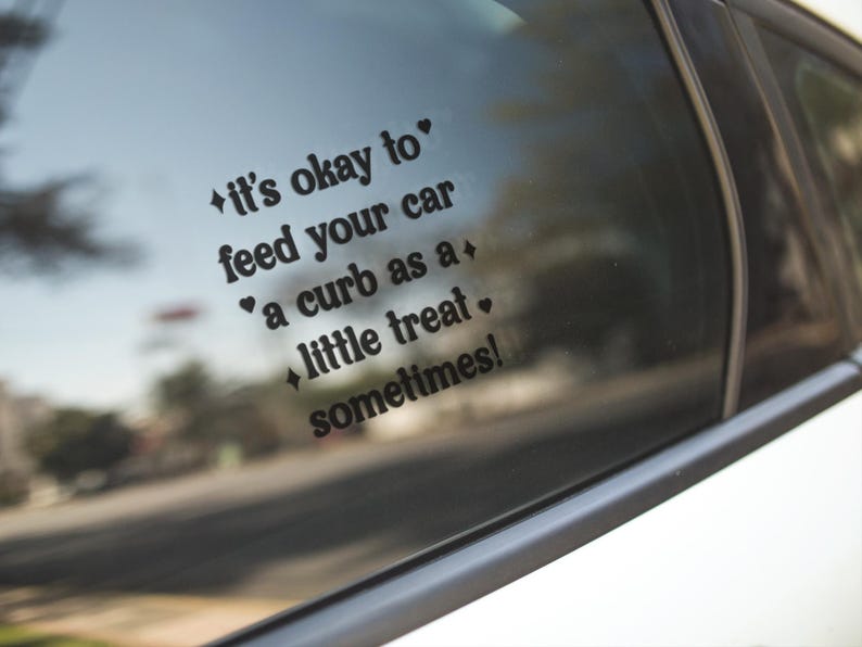 May include: A black vinyl decal applied to a car window featuring the text 'it's okay to feed your car a curb as a little treat sometimes!' in a bold, rounded font, accented with small heart and four-pointed star symbols.