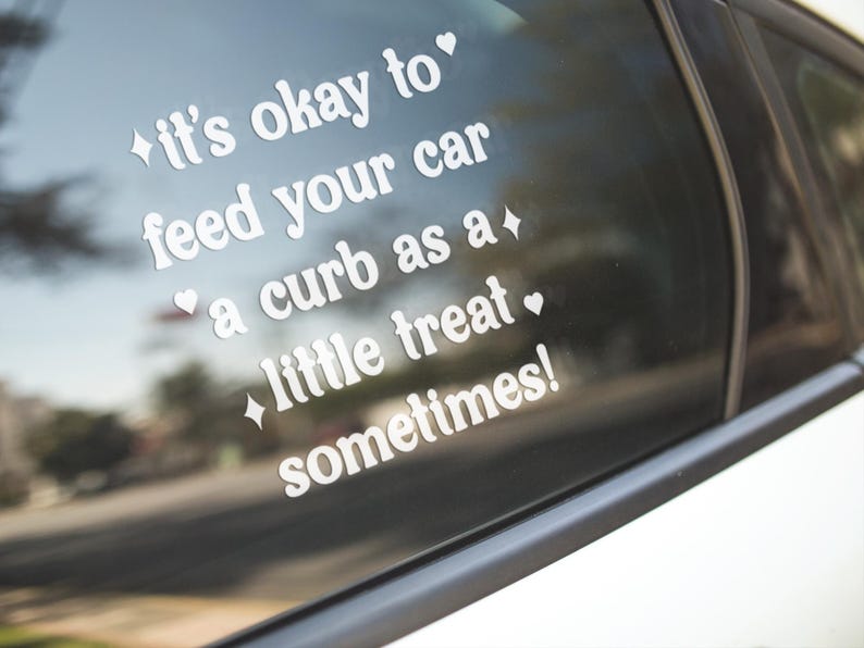 May include: A white vinyl decal applied to a car window featuring the text "it's okay to feed your car a curb as a little treat sometimes!" written in a rounded lowercase font, accented with small star and heart symbols surrounding the text.