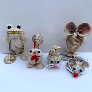 May include: A collection of four animal figurines crafted from various seashells. The set includes a frog, a chicken, a mouse, and an owl, all featuring googly eyes. The figures are constructed from ribbed, brown and white shells with added details like red felt accents and white ribbon whiskers.