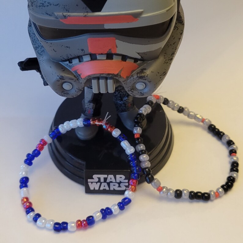 Pu&ograve; includere: Due braccialetti di perline, uno con perline bianche, blu e rosse, e l'altro con perline nere, bianche e grigie. I braccialetti sono su una superficie nera con la scritta "STAR WARS" stampata.