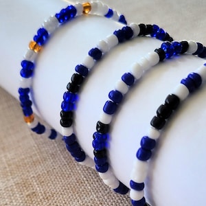 Peut inclure: Trois bracelets en perles blanches, bleues et noires. Les bracelets sont superposés, le bracelet blanc étant sur le dessus. Les bracelets sont faits de petites perles rondes.