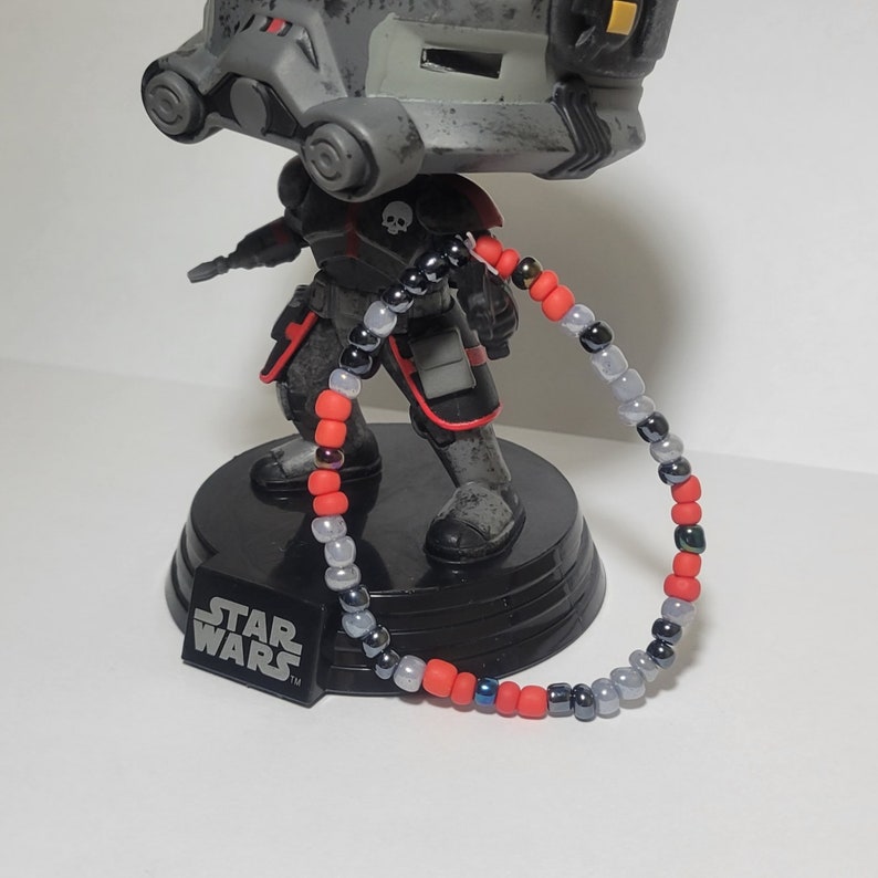 Puede incluir: Un brazalete de cuentas rojas, negras y plateadas con un logotipo de Star Wars en la base de una figura.