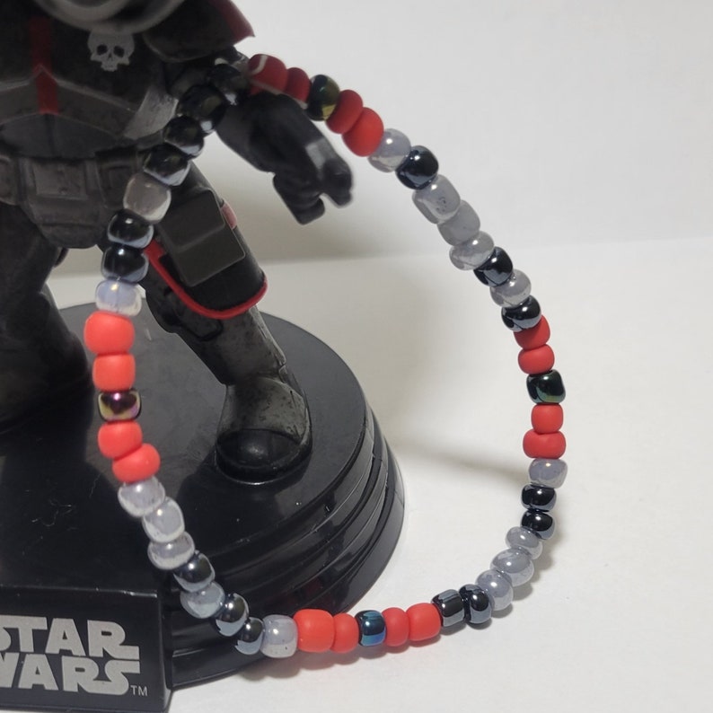 Puede incluir: Un brazalete de cuentas rojas, grises y negras. Las cuentas son peque&ntilde;as y redondas. El brazalete est&aacute; hecho de una variedad de materiales, incluyendo vidrio, metal y pl&aacute;stico.