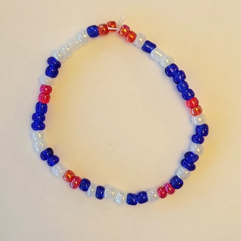 Pu&ograve; includere: Un braccialetto di perline bianche, blu e rosse. Le perline sono disposte in un motivo ripetitivo.
