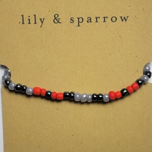 Puede incluir: Primer plano de una pulsera de cuentas con cuentas negras, grises y rojas alternadas. La pulsera est&aacute; sobre una tarjeta marr&oacute;n con el texto ".lily & sparrow" impreso en ella.