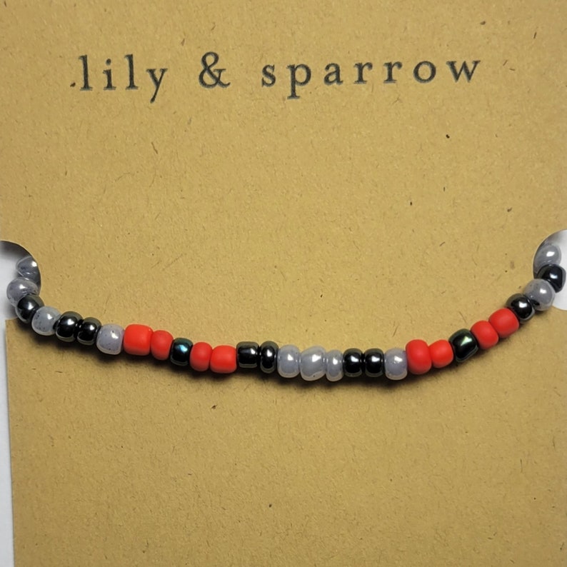 Puede incluir: Primer plano de una pulsera de cuentas con cuentas negras, grises y rojas alternadas. La pulsera est&aacute; sobre una tarjeta marr&oacute;n con el texto ".lily & sparrow" impreso en ella.