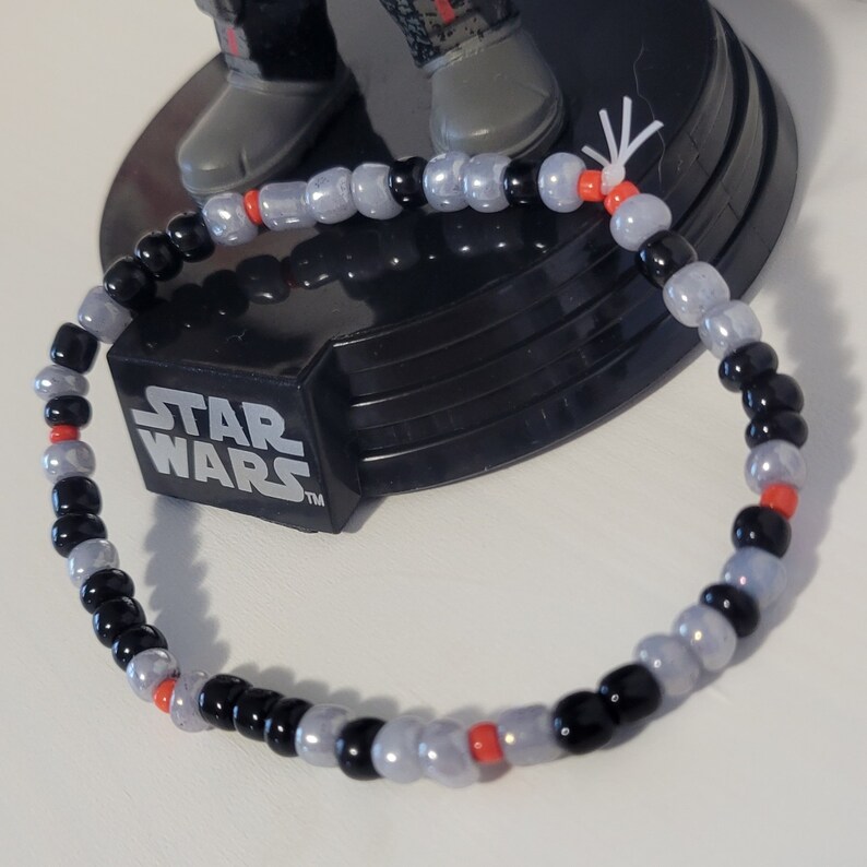 Pu&ograve; includere: Un braccialetto di perline nere e grigie con accenti rossi. Il braccialetto &egrave; realizzato con piccole perline rotonde e ha un design semplice.