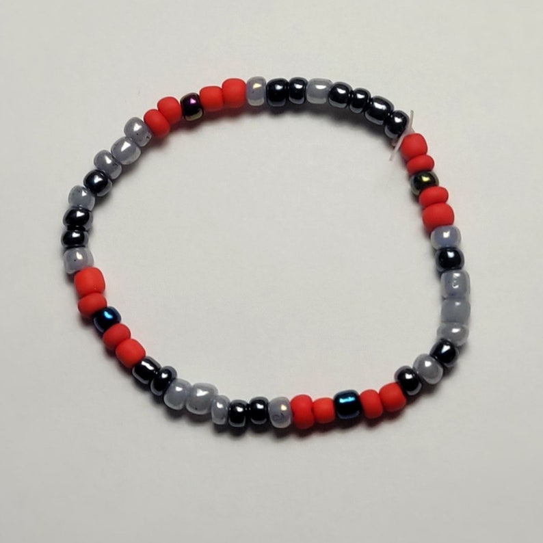 Puede incluir: Un brazalete de cuentas rojas, grises y negras. El brazalete est&aacute; hecho con cuentas peque&ntilde;as y redondas.