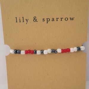 May include: A red, white and black beaded bracelet with the text "lily & sparrow" on a brown card.