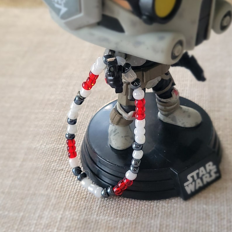 May include: A white, red and black beaded bracelet with a silver charm. The bracelet is wrapped around the leg of a Star Wars action figure.