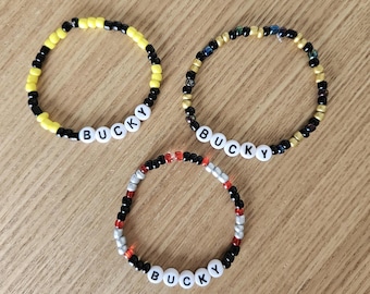 Puede incluir: Tres pulseras de cuentas con el nombre "BUCKY" escrito con cuentas de letras blancas. Las pulseras presentan cuentas negras, amarillas, doradas, rojas y plateadas. Las pulseras están sobre una superficie de madera marrón claro.