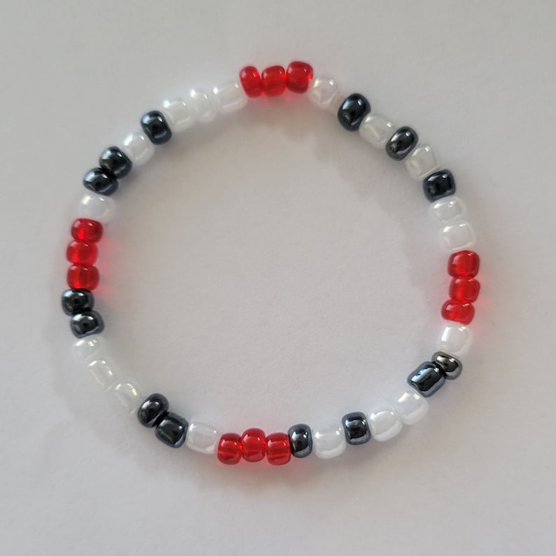 May include: A white, red and black beaded bracelet. The bracelet is made with small round beads and has a simple design.