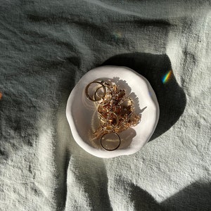 Könnte beinhalten: Eine weiße, organisch geformte Keramikschale mit einer Auswahl an goldenen Ringen und Schmuck. Die Schale liegt auf einem strukturierten, hellgrünen Stoff, mit Schatten und einem Regenbogen-Prisma-Effekt.