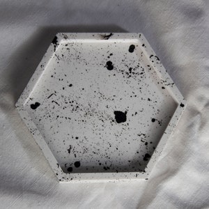 Könnte beinhalten: Ein weißes sechseckiges Tablett mit schwarzen Spritzflecken. Das Tablett ist aus Beton und hat eine glatte, polierte Oberfläche.