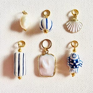 Könnte beinhalten: Ein Set aus sechs goldenen Charms mit weißen und blauen Porzellan-Designs. Die Charms umfassen eine Perle, eine gestreifte Kugel, eine Muschel, ein gestreiftes Rechteck, ein rosafarbenes Rechteck mit einem weißen Design und einen gestreiften Fisch.