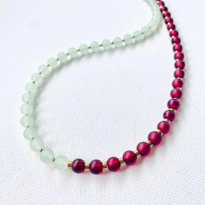 Può includere: Una collana di perline con due sezioni distinte. Un lato ha perline verde chiaro, l'altro ha perline rosso intenso. Distanziatori color oro separano le perline. La collana è esposta su una superficie bianca.