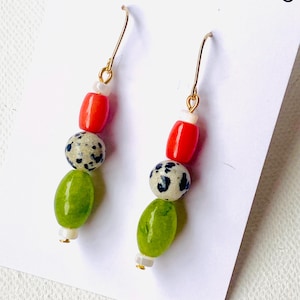 May include: Dangle earrings with gold-coloured hooks. Each earring has a small white bead, a red barrel bead, a speckled bead, a green oval bead, and a small white bead. The earrings are displayed on a white card.