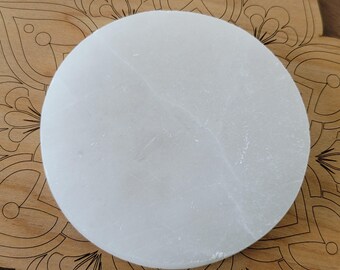 May include: A round, white selenite charging plate. The plate is smooth and sits on a wooden surface with a decorative mandala design. The selenite has natural variations in its surface.
