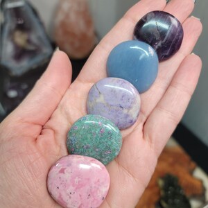 May include: A hand holds five polished, round gemstones of various colors. The stones include pink, green, purple, blue, and a dark purple stone. The gemstones are smooth and shiny, with visible patterns and textures.